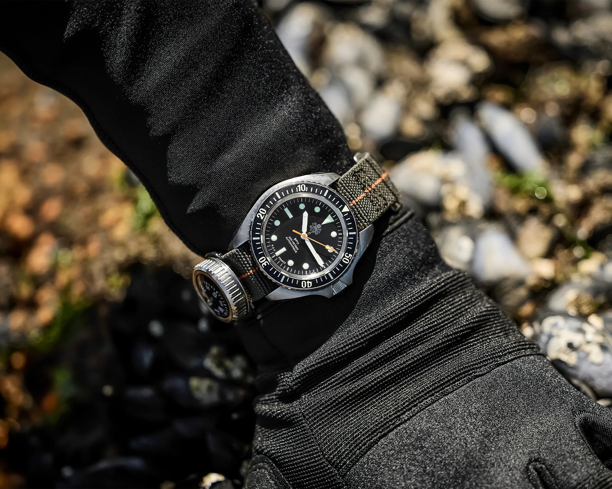 Black dive watch with a rotating bezel and green fabric strap worn on a wrist in a black wetsuit.