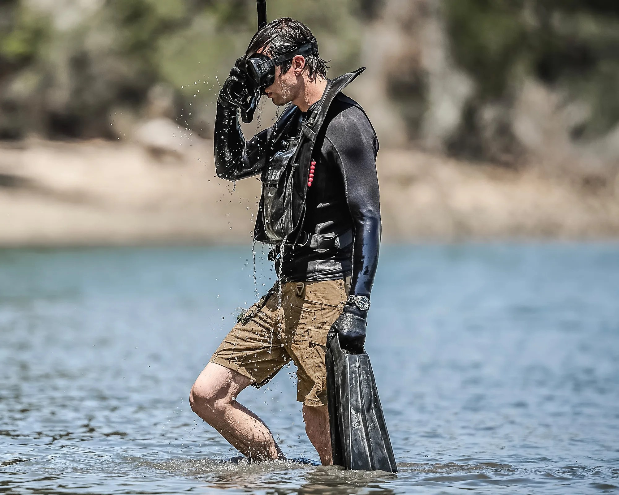 Person in black wetsuit top and brown shorts holding black diving fins and adjusting black diving mask while standing in shallow water.