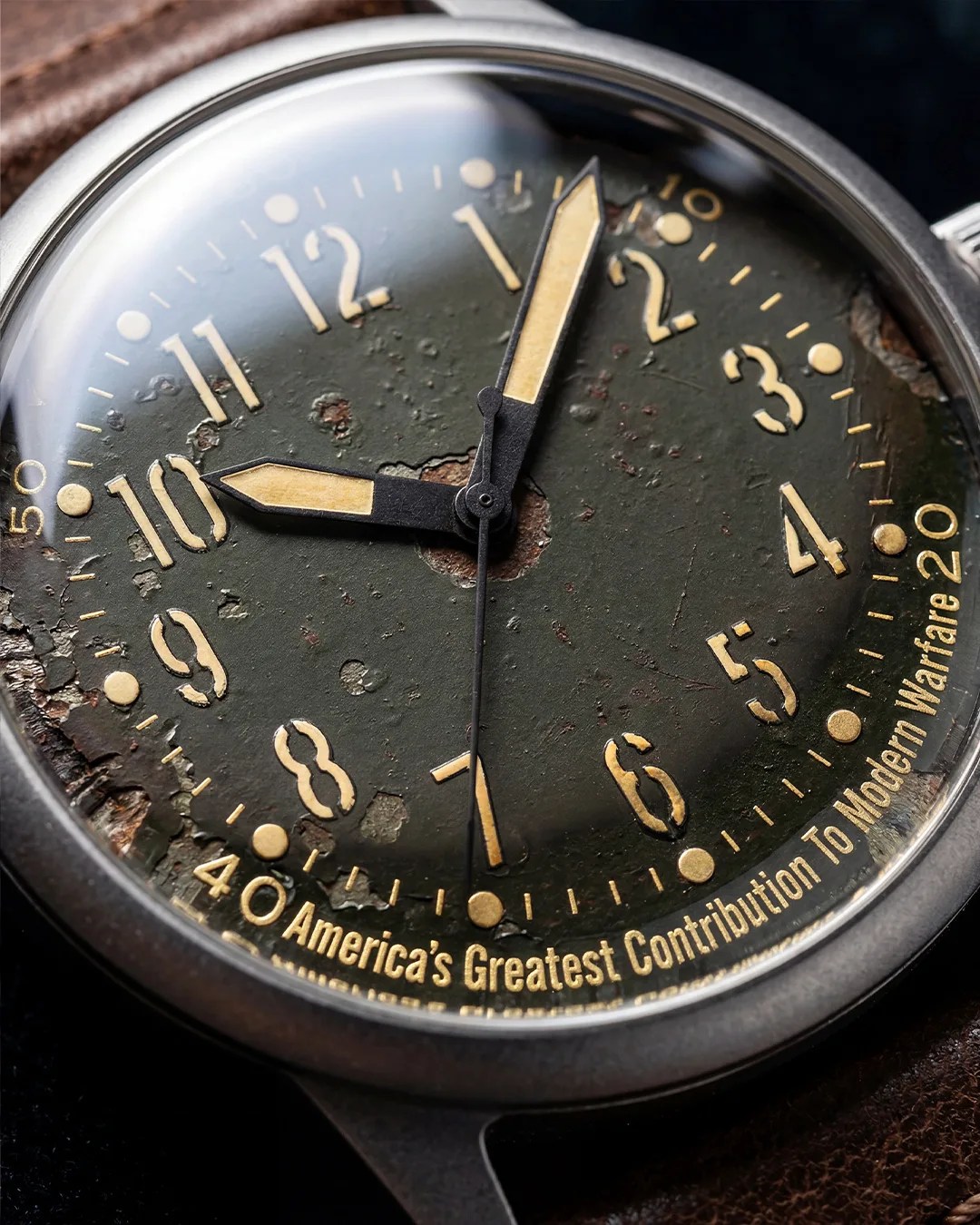 Close-up of a vintage watch face with worn black dial, yellowed numerals, and text reading "America's Greatest Contribution To Modern Warfare 20".