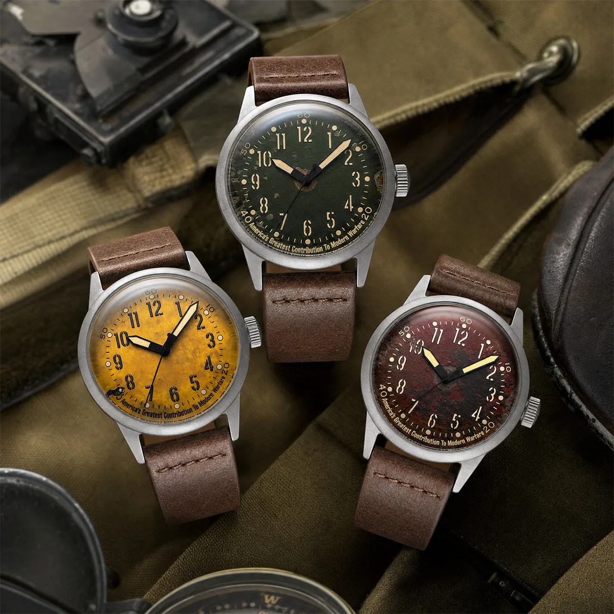 Three vintage-style wristwatches with brown leather straps and distressed yellow, green, and red dials on a fabric background.