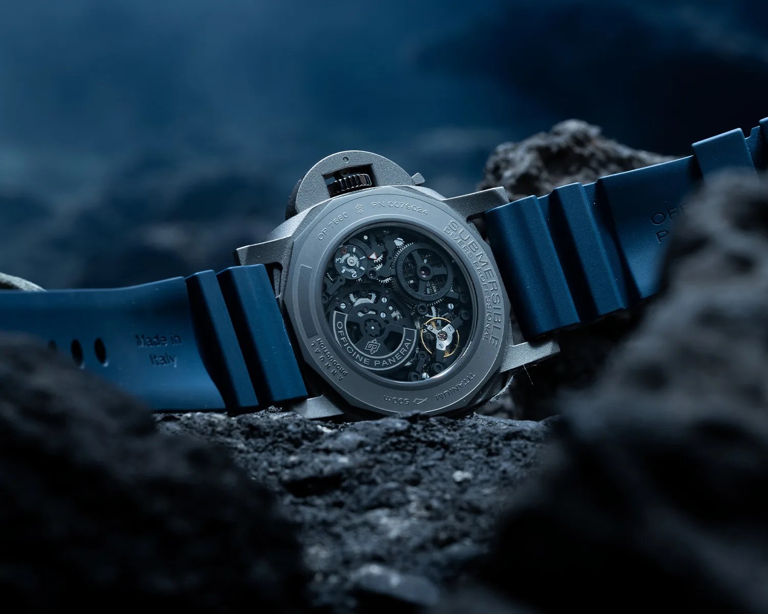 Titanium Officine Panerai Submersible watch with visible mechanical movement and blue rubber strap on dark rocky surface.