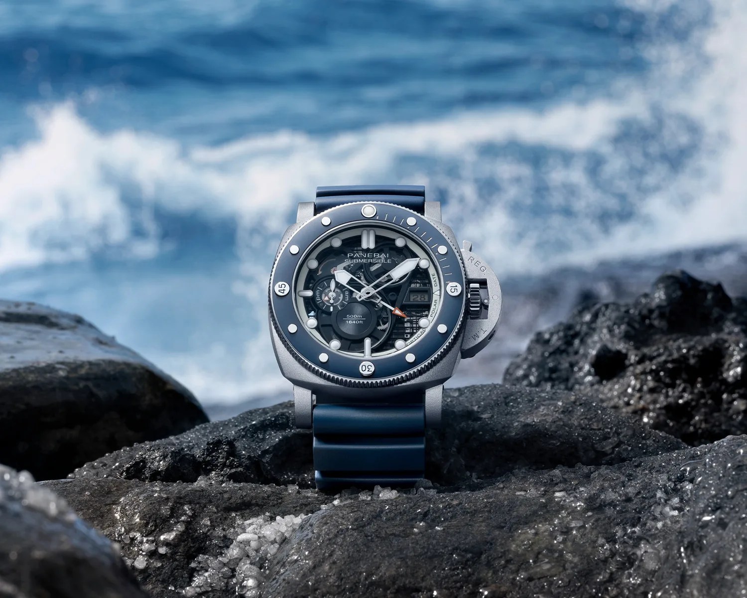 Panerai Submersible dive watch with blue bezel and strap on dark volcanic rocks by the ocean.