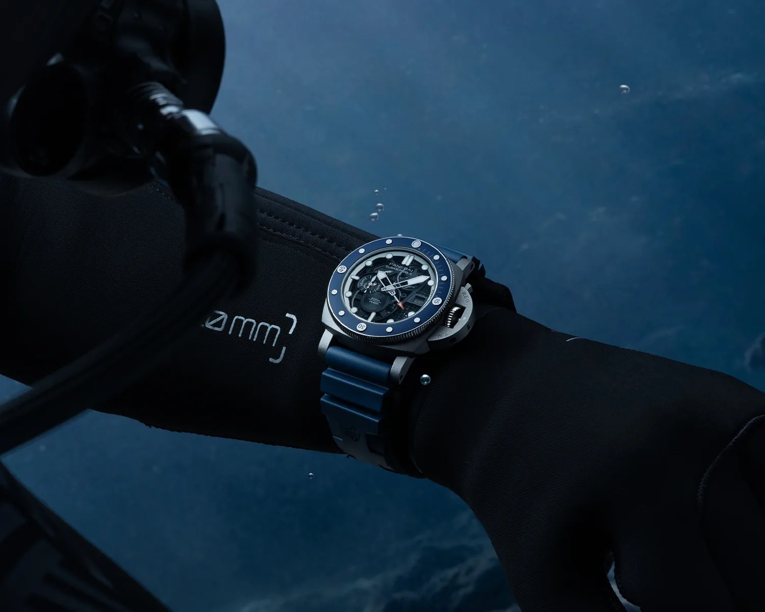 Diver's wristwatch with blue bezel and strap worn over a black wetsuit sleeve underwater.