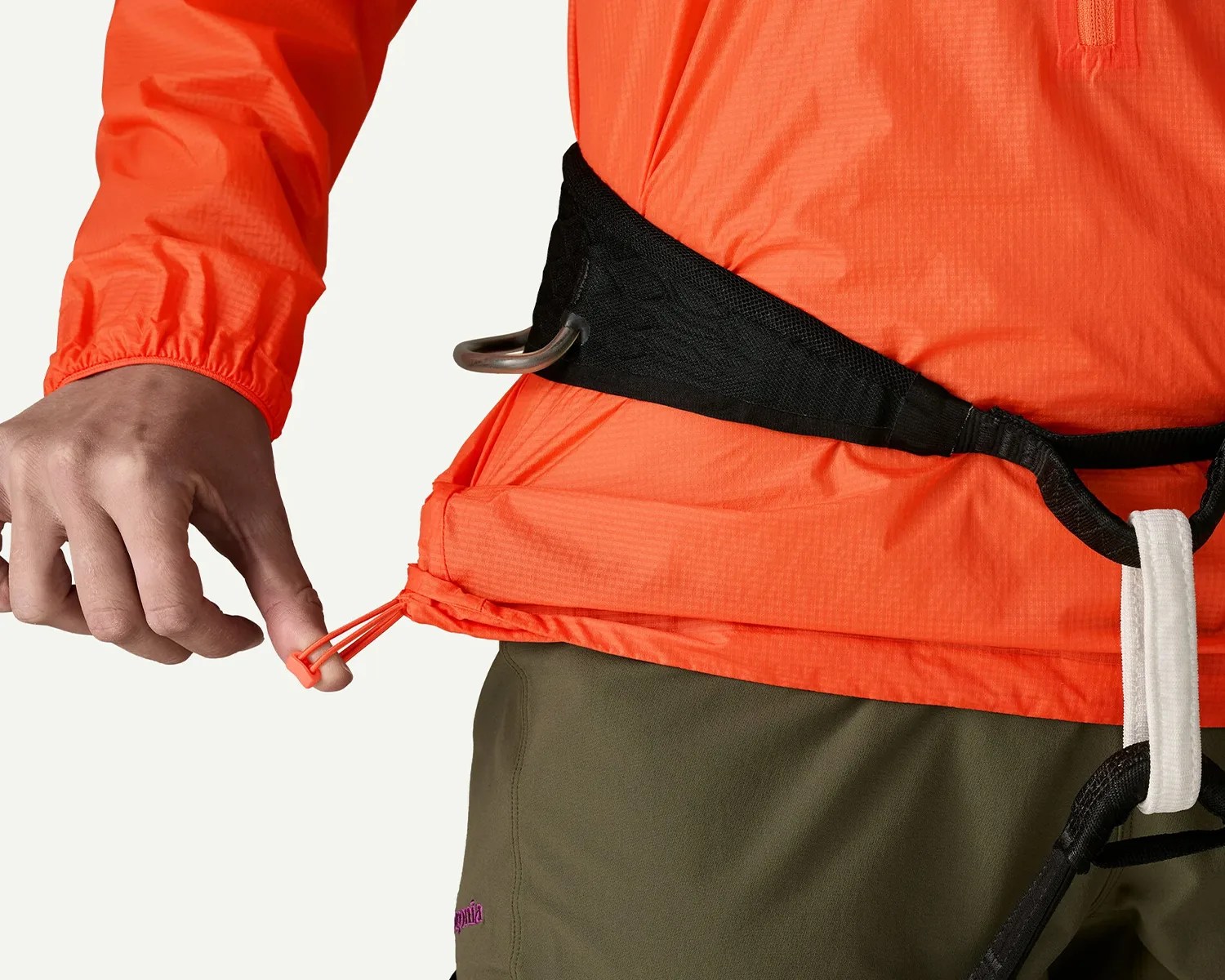 Close-up of a person adjusting the orange drawcord hem of an orange jacket while wearing a black climbing harness and olive pants.
