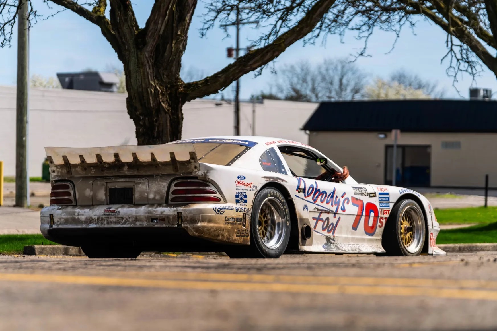 White and dirty vintage race car with "Nobody's Fool" and number 70 decals parked under a tree.