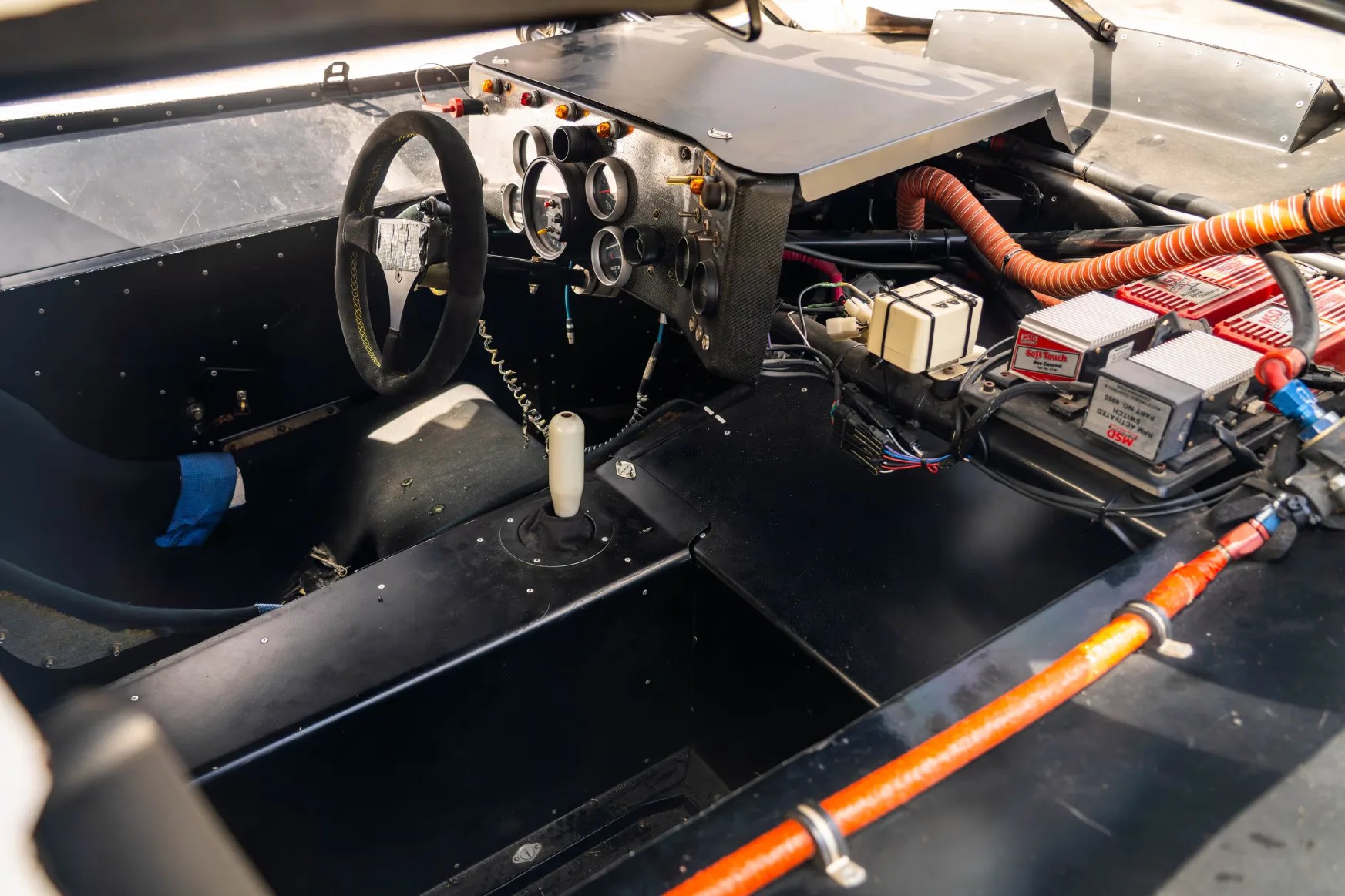 Minimalist black race car cockpit with suede steering wheel, white gear shifter, analog gauges, and exposed wiring.