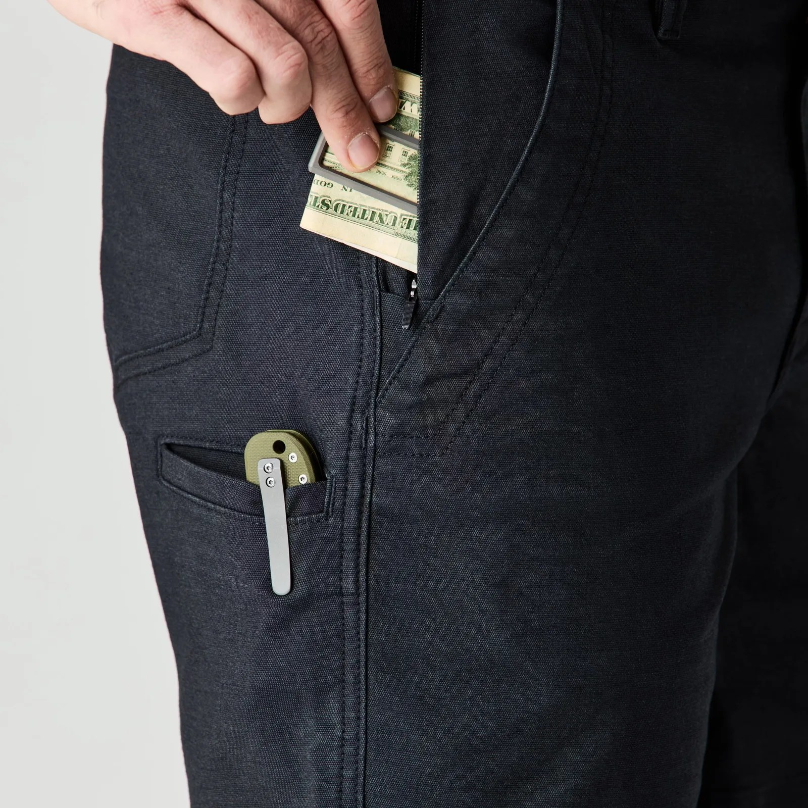 Hand placing folded US dollar bills into a zippered pocket of black pants with a green-handled folding knife clipped to a smaller pocket.