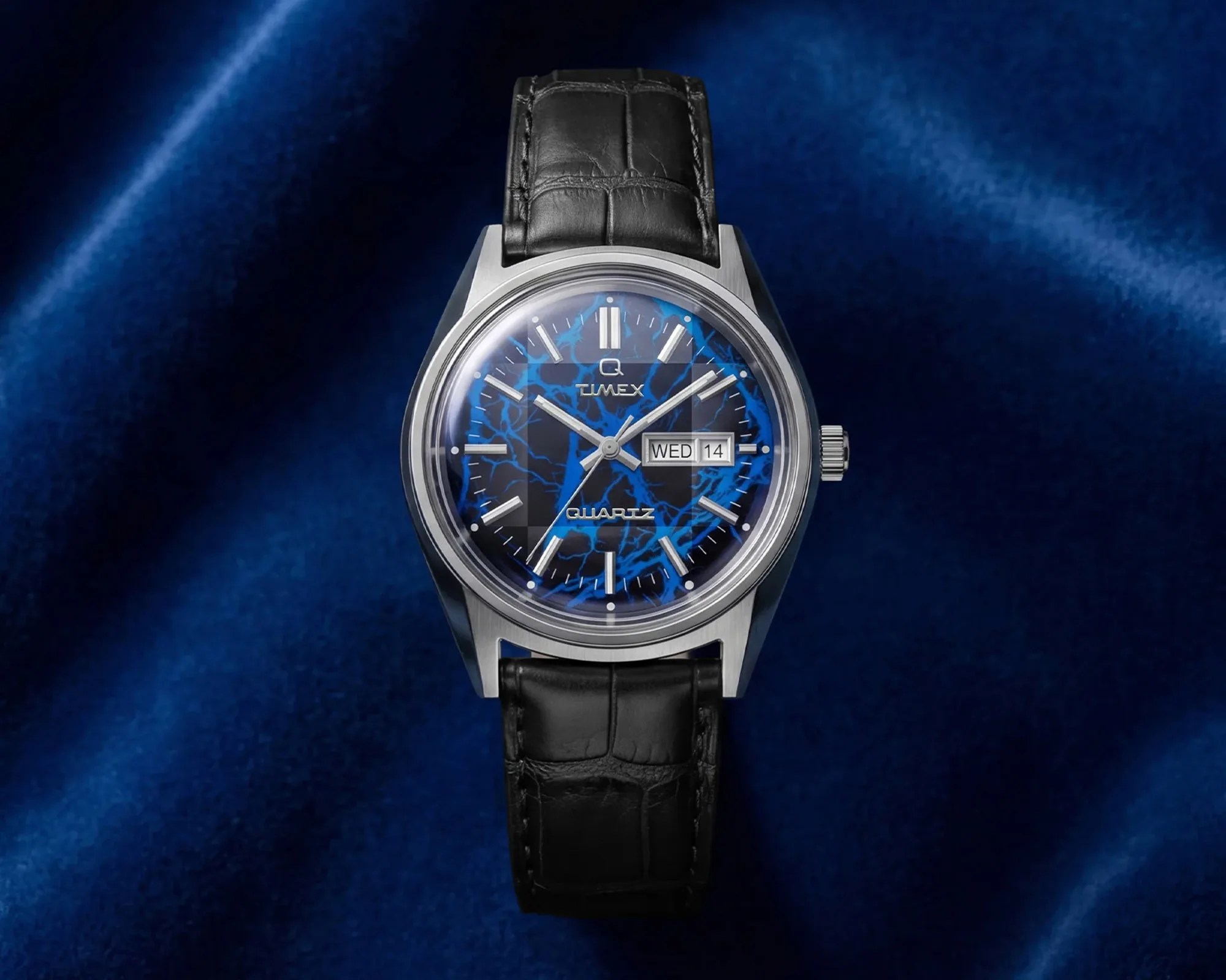 Silver Timex wristwatch with black leather strap and blue lightning-patterned dial showing day and date.