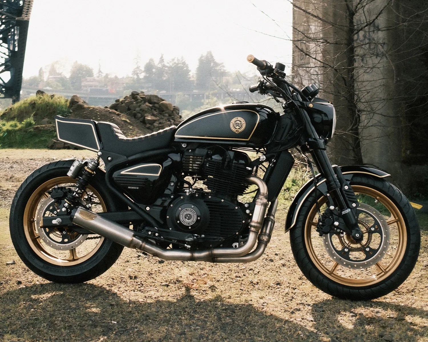 Black and gold Royal Enfield motorcycle with a custom exhaust and cafe racer style seat parked on gravel.