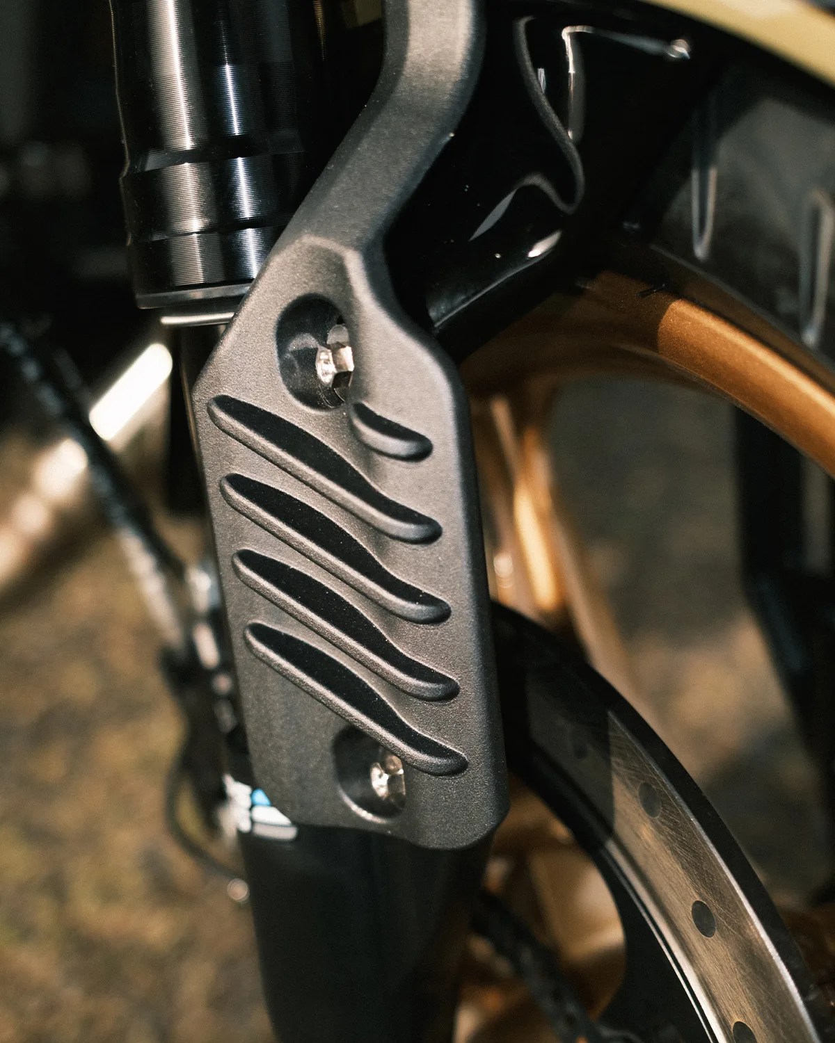 Close-up of a black textured metal bike fork guard attached with two silver bolts near a gold-colored wheel rim and disc brake rotor.
