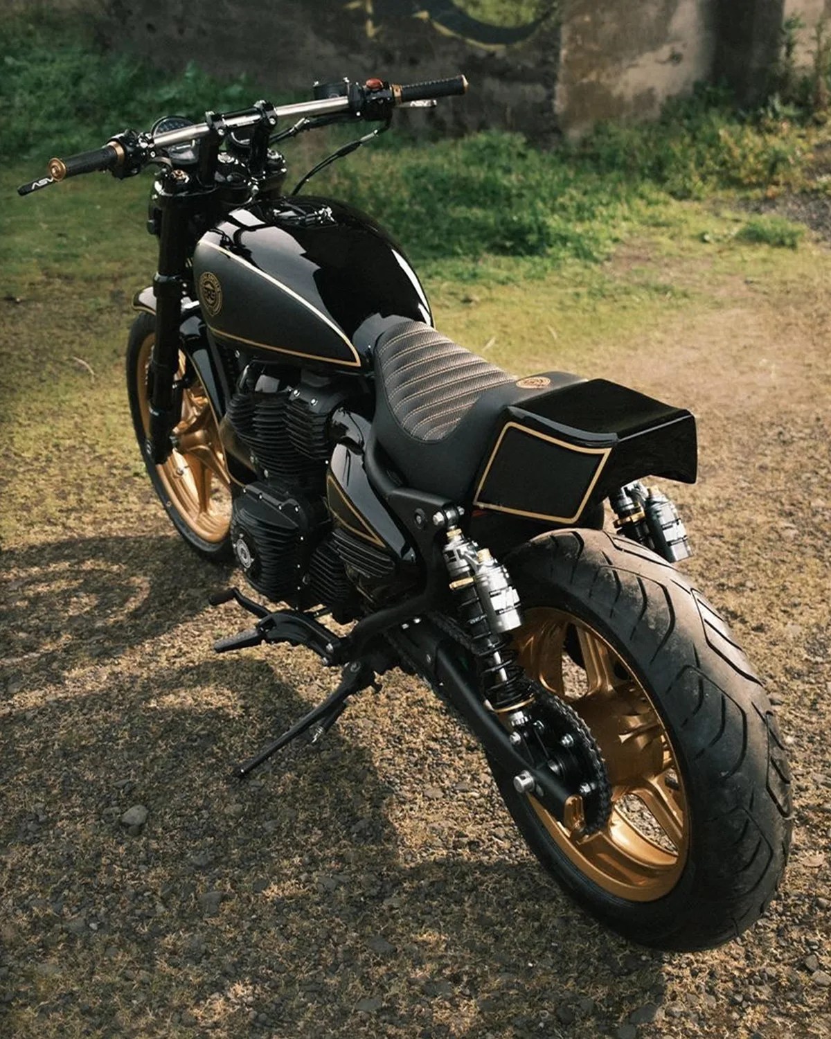 Black and gold custom motorcycle with quilted seat and dual rear shock absorbers on gravel ground.