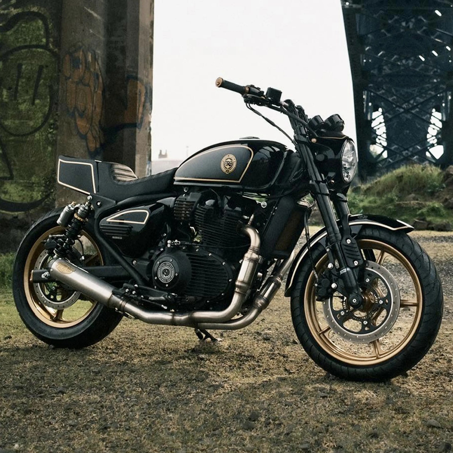 Black and gold Royal Enfield motorcycle parked on gravel under a bridge with graffiti on a pillar.