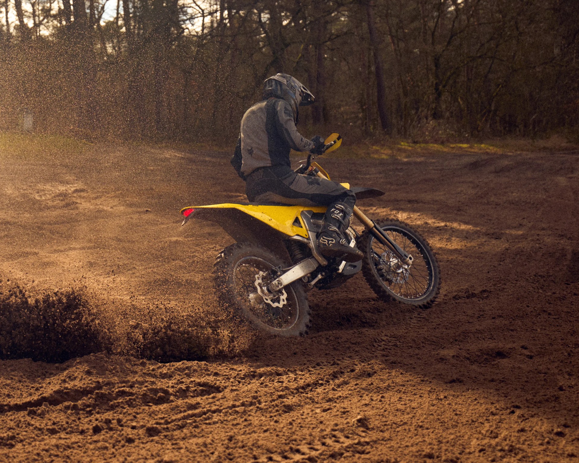 Person wearing gray and black gear riding a yellow dirt bike on a muddy trail with dirt spraying behind.