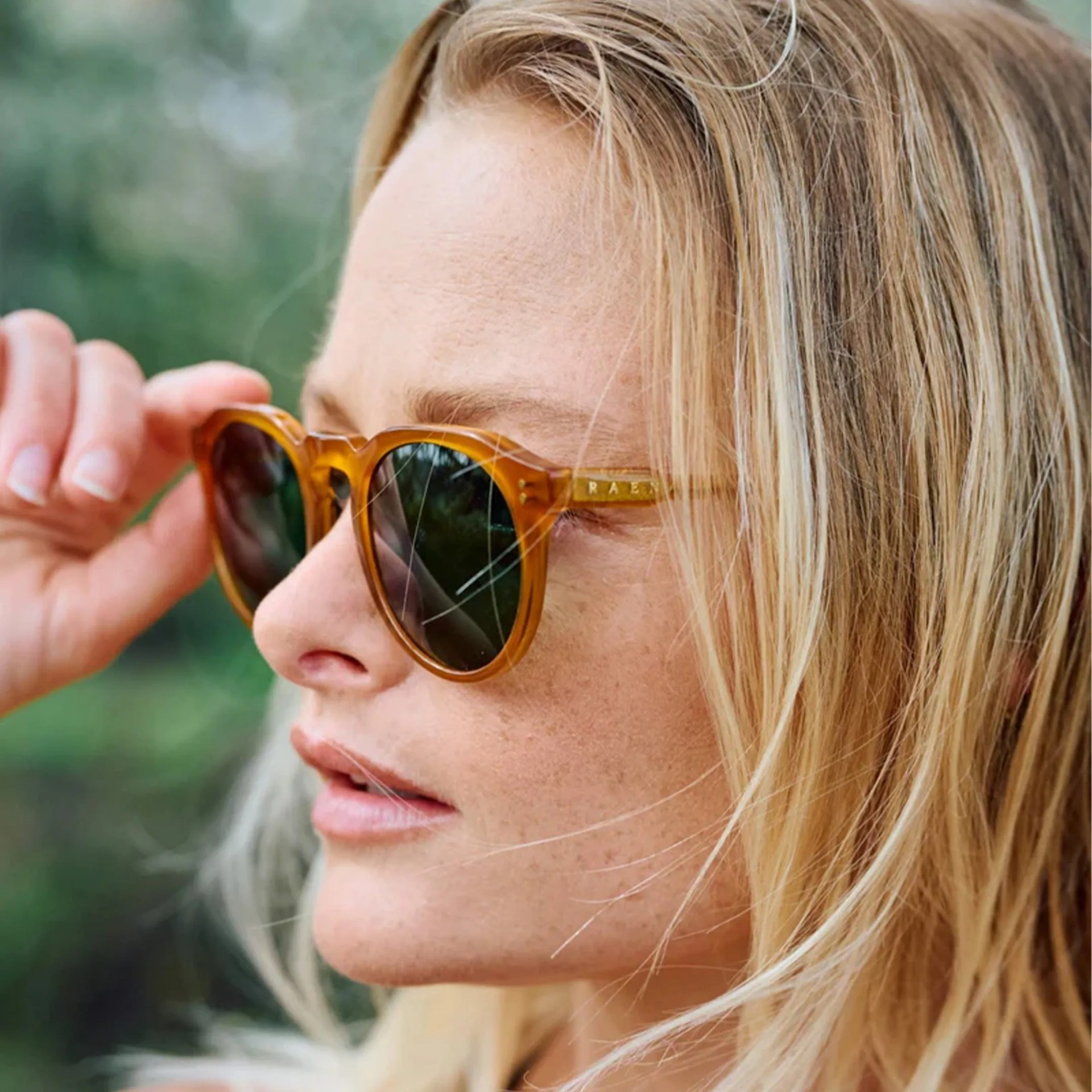 Close-up of a person with blonde hair adjusting amber-colored sunglasses with dark lenses.