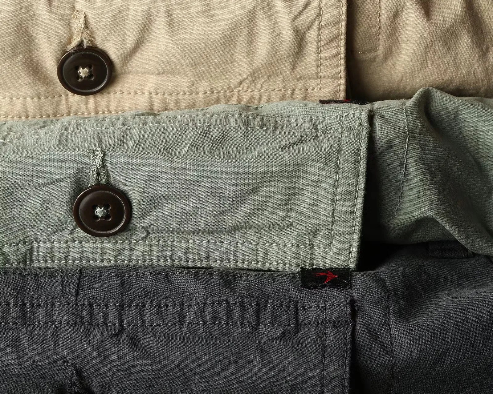 Close-up of three folded shirts in beige, green, and dark gray, each with a visible button and stitching details.