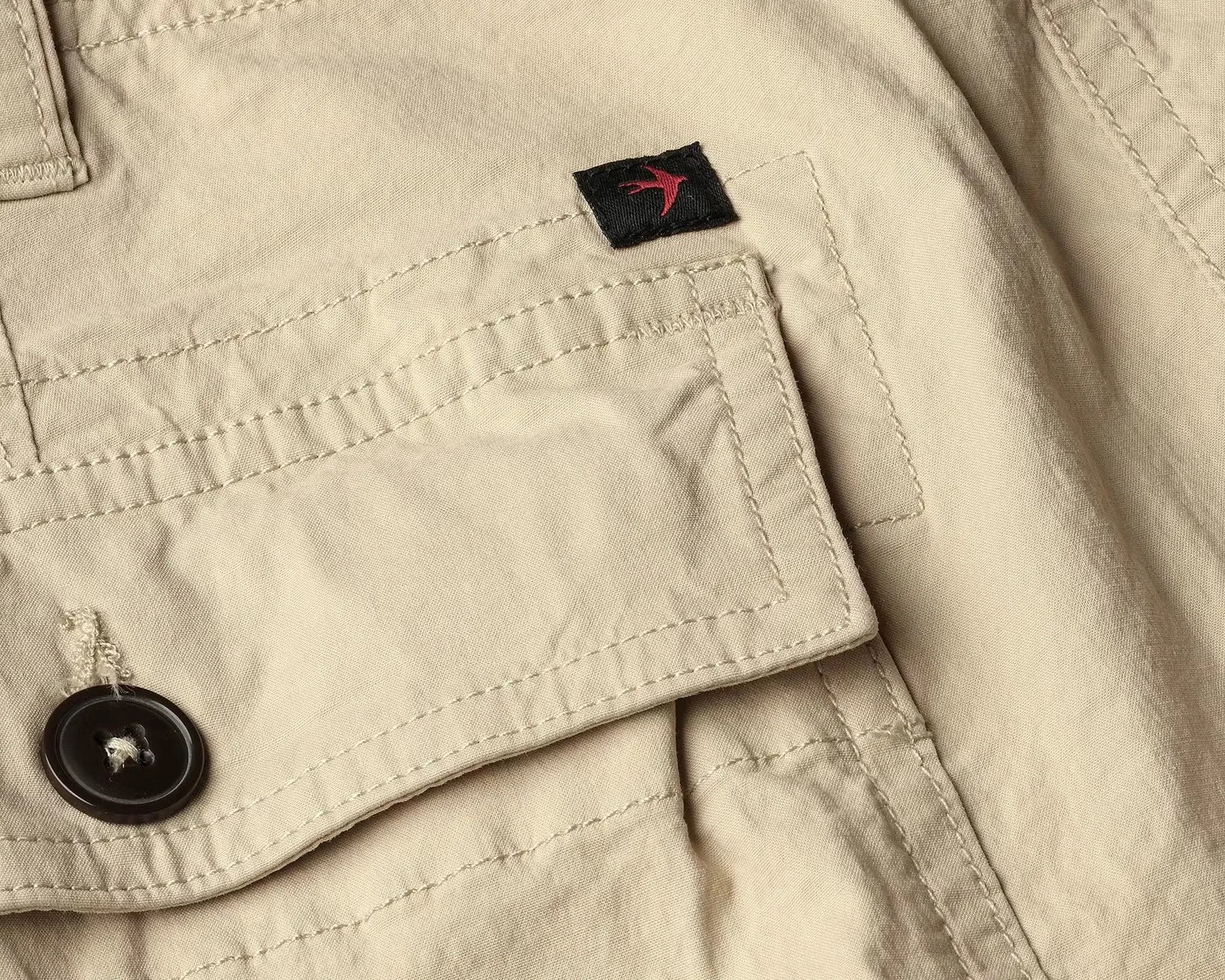 Close-up of beige fabric with detailed stitching, a black button, and a small black tag featuring a red bird logo.