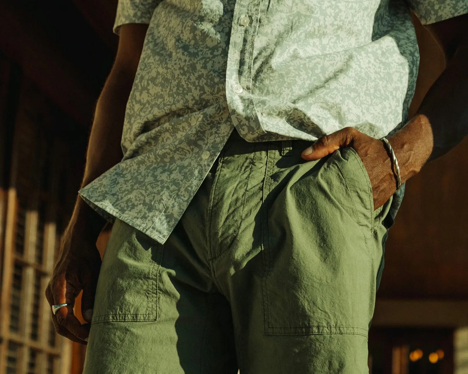 Close-up of a person wearing a light patterned button-up shirt and olive green pants with one hand in the pocket and silver jewelry.