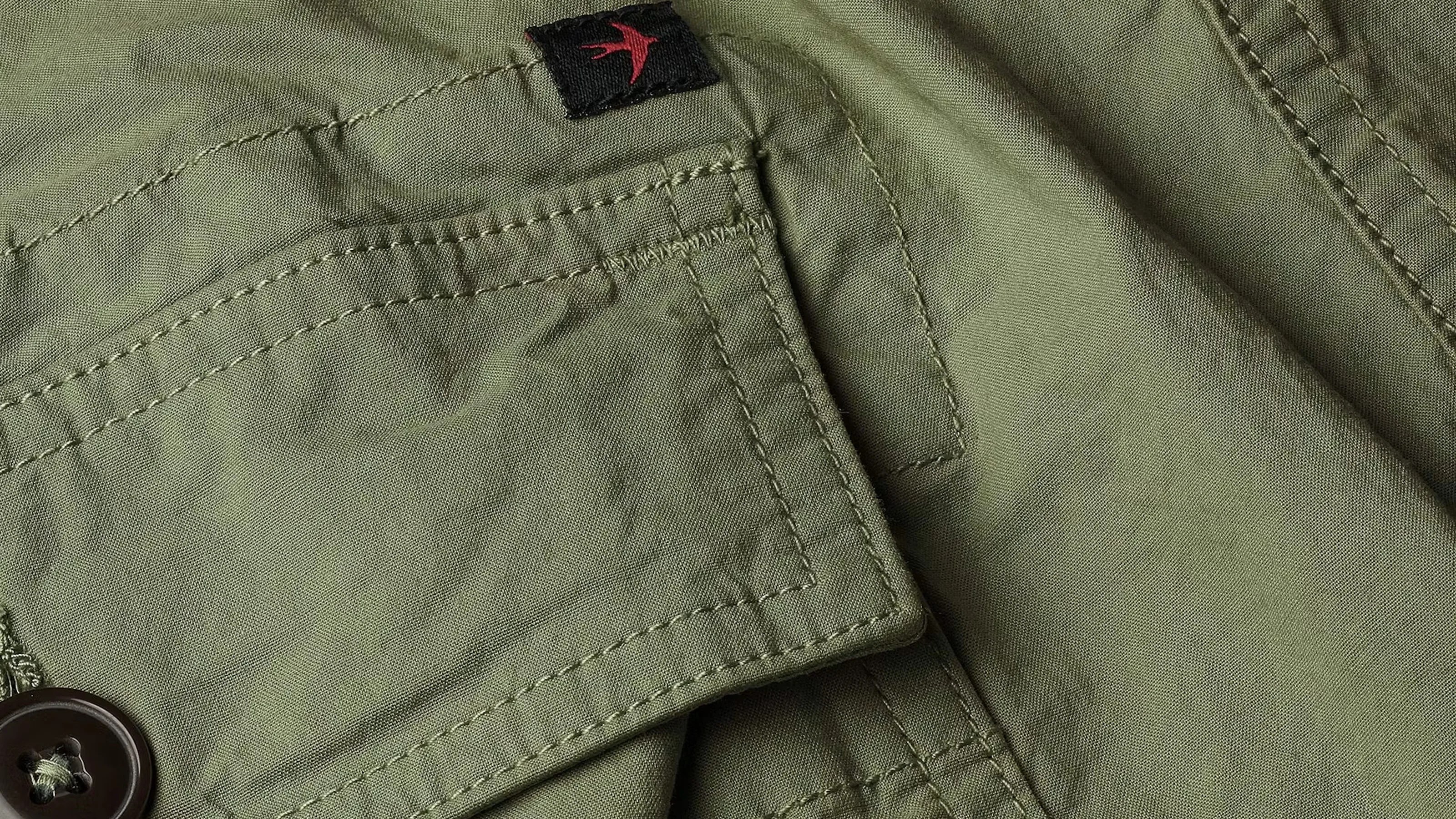 Close-up of olive green fabric with detailed stitching, a button, and a small black tag with a red bird logo.
