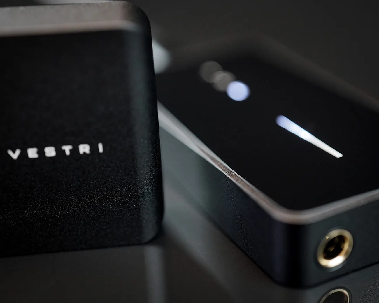 Black rectangular electronic device with illuminated indicators and a gold headphone jack next to a black box with "VESTRI" text.