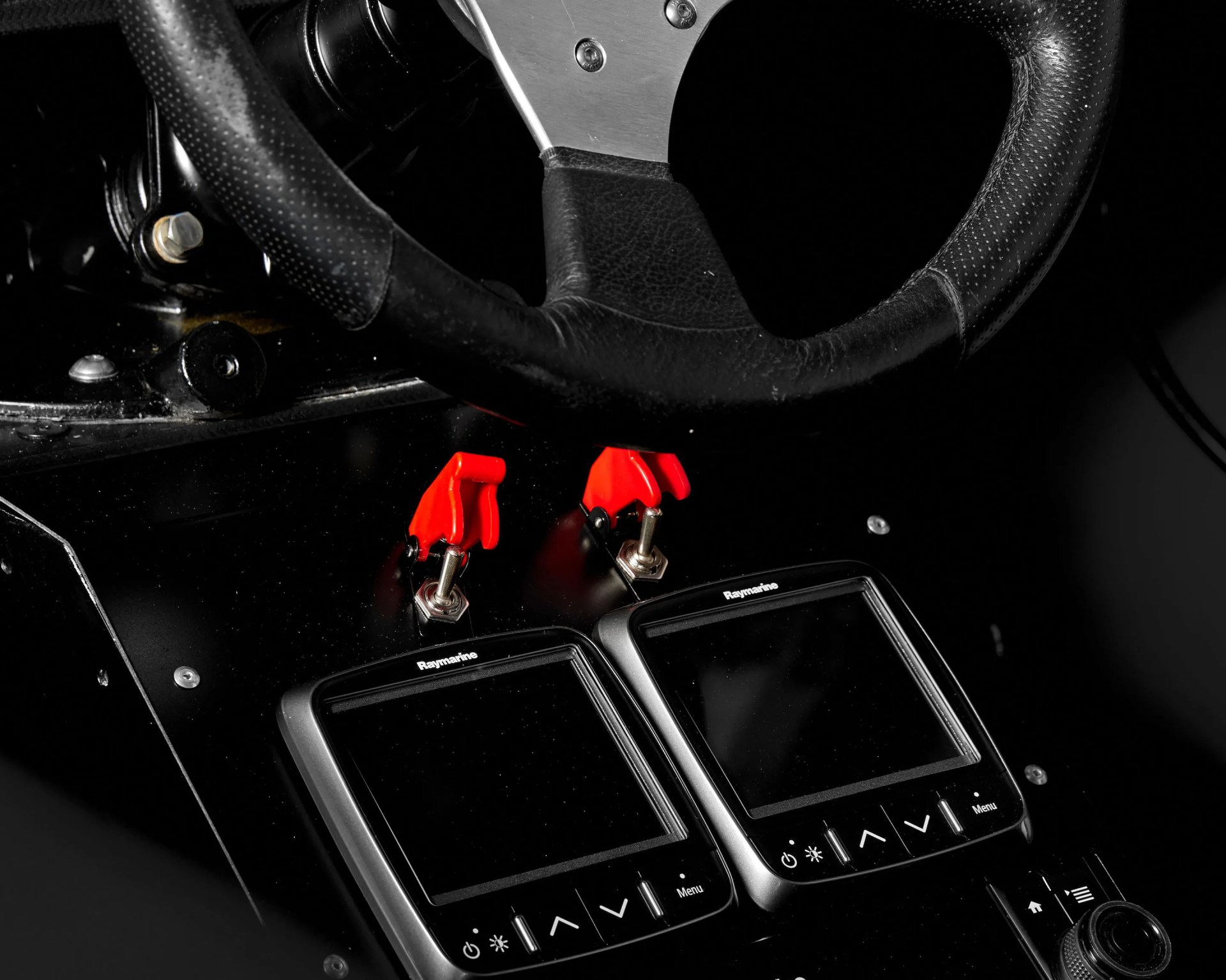 Close-up of a black leather steering wheel above two Raymarine marine navigation screens with red toggle switches.