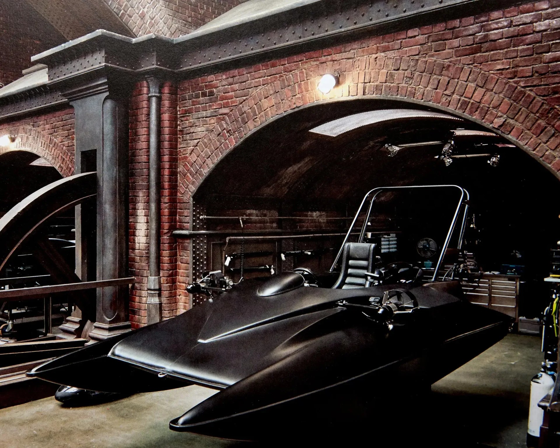 Matte black Batmobile with sleek angular design parked inside a brick-walled garage with industrial lighting.