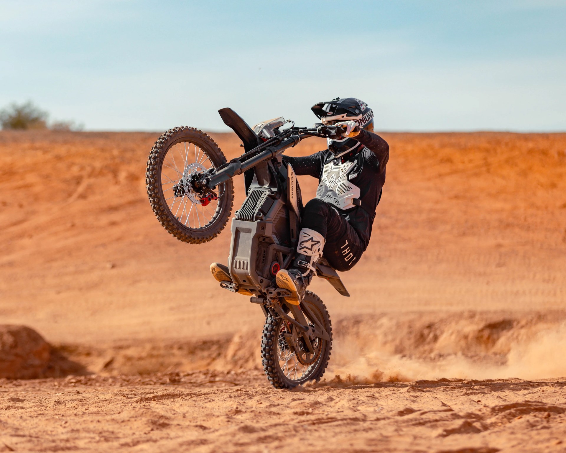 Person wearing black and white protective gear performing a wheelie on a black off-road motorcycle in a desert.