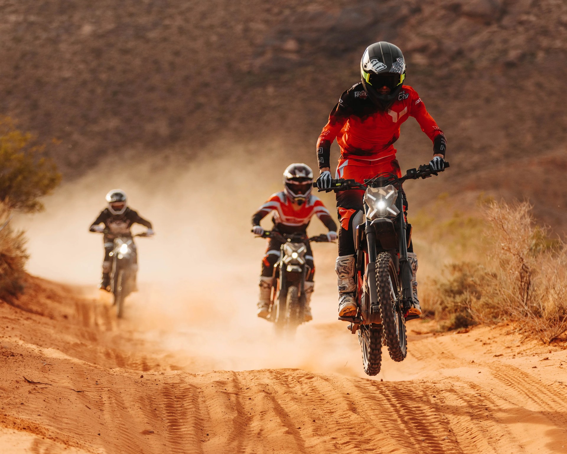Three motocross riders in helmets and gear riding on a dusty dirt trail in a desert landscape.