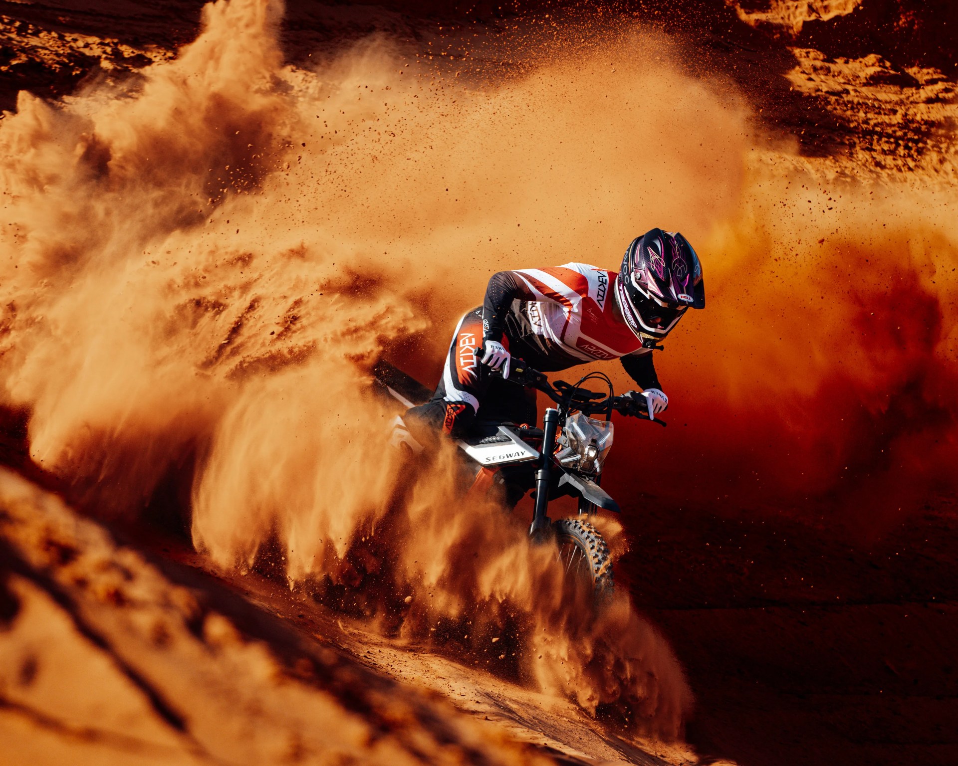 Motorcyclist in black and orange gear kicking up a large cloud of orange dust while riding a Segway dirt bike.