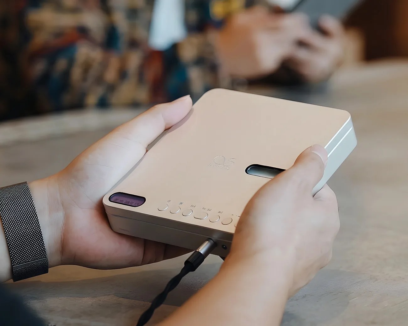 Person holding a beige Q5 SoundPro audio device with buttons and a connected cable on a table.