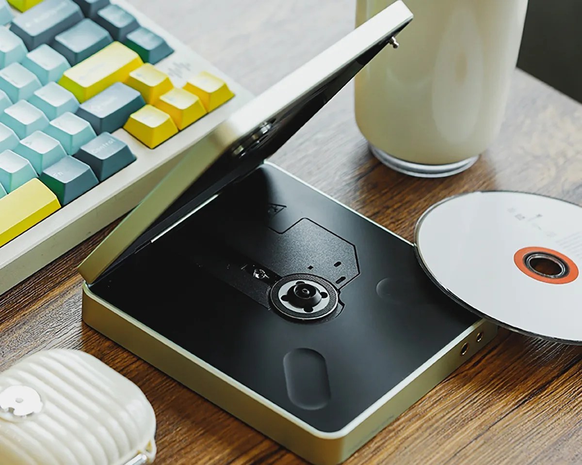 External CD/DVD drive with an open lid and a disc partially inserted on a wooden desk near a colorful keyboard and a white mug.