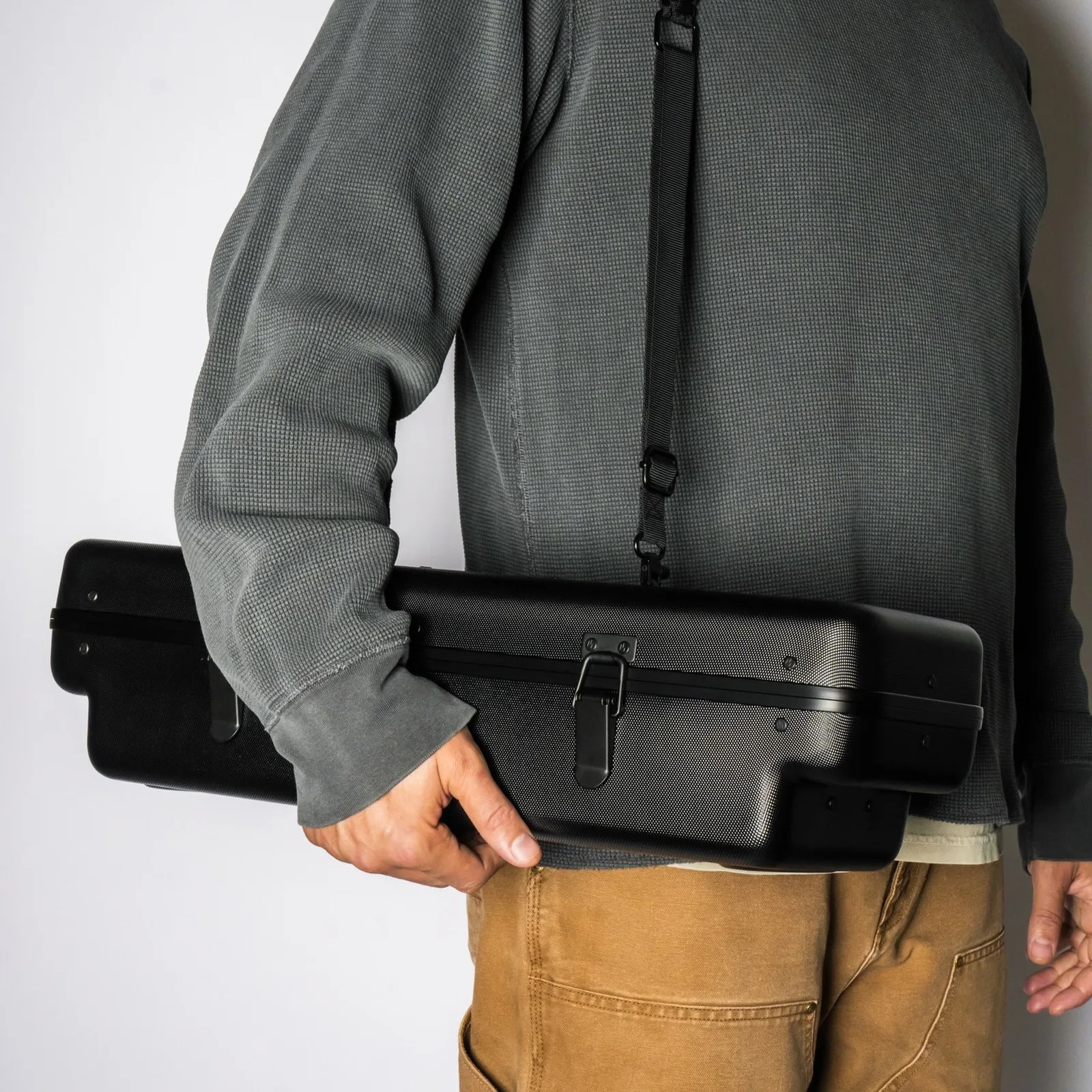 Person wearing a gray textured long-sleeve shirt and brown pants holding a black hard case with a shoulder strap.