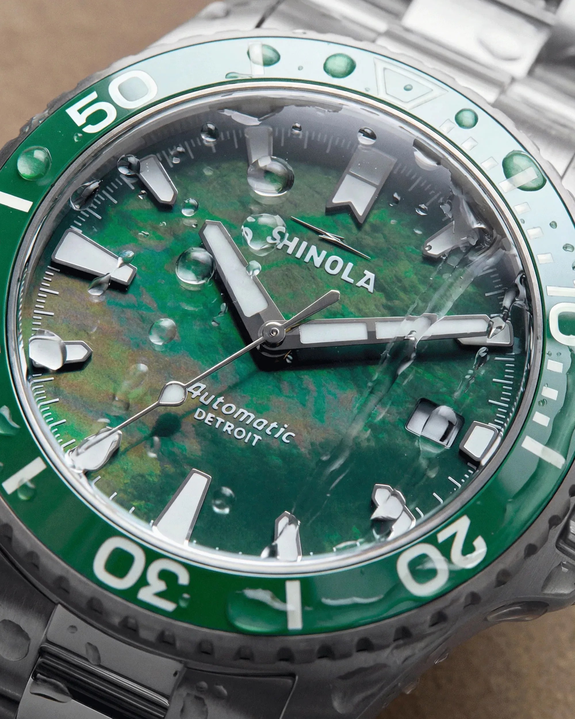 Close-up of a Shinola automatic watch with a green dial and bezel, water droplets on the glass, and a stainless steel bracelet.