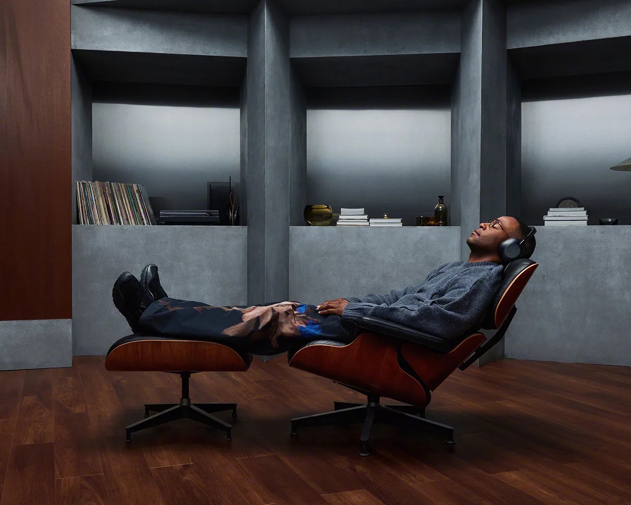 Man reclining on a mid-century modern wooden lounge chair with matching ottoman, wearing headphones in a room with gray walls and wooden floor.
