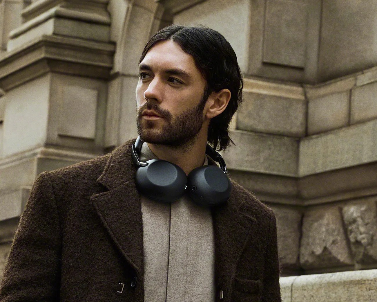 Man wearing black over-ear headphones around his neck and a brown textured coat in an urban stone building setting.