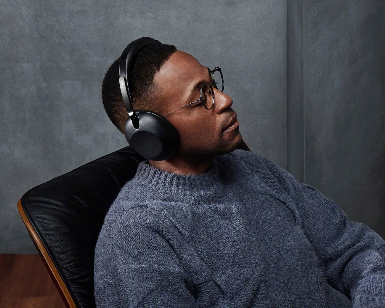 Man wearing black over-ear Sony headphones and round glasses, sitting in a black leather chair against a gray wall.