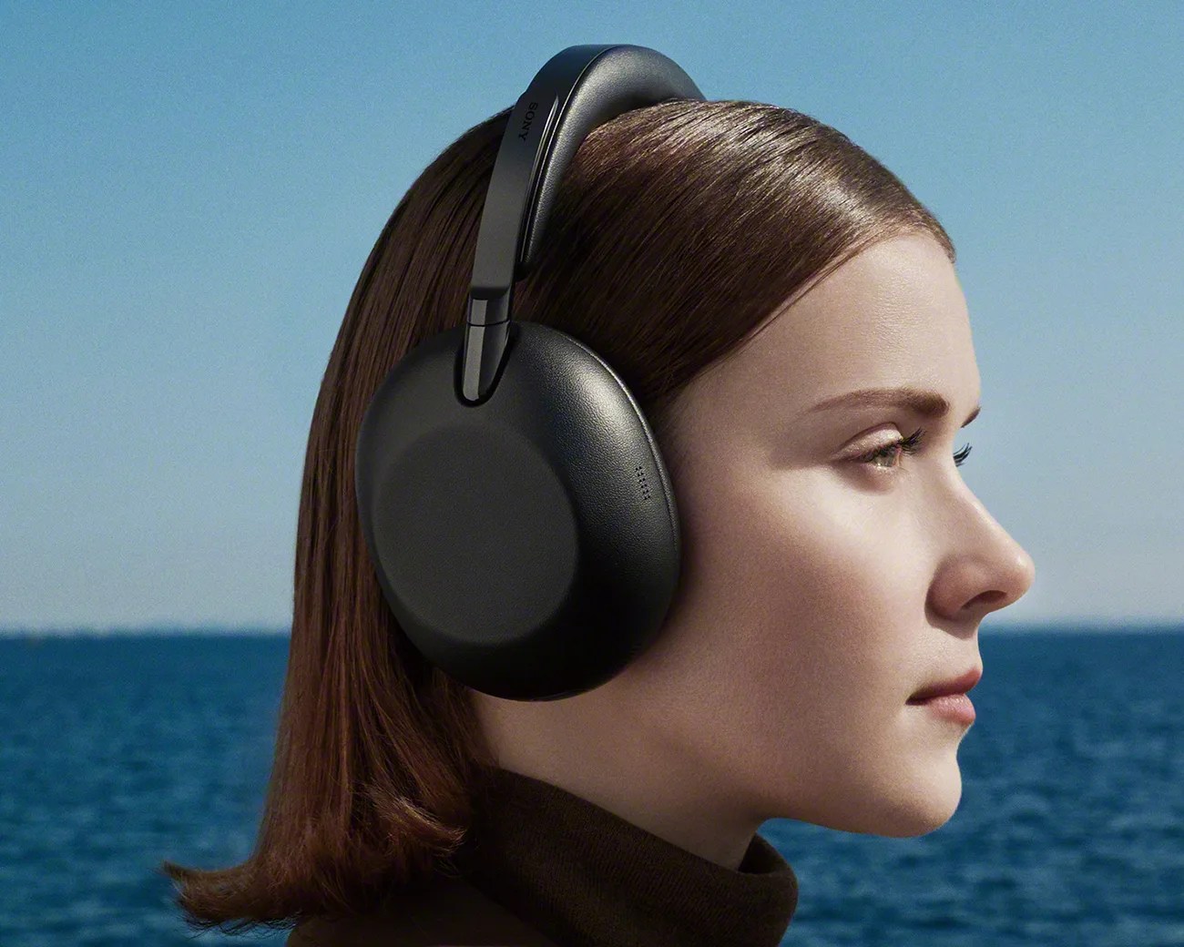 Black over-ear Sony headphones worn by a person with brown hair against a sea and sky background.