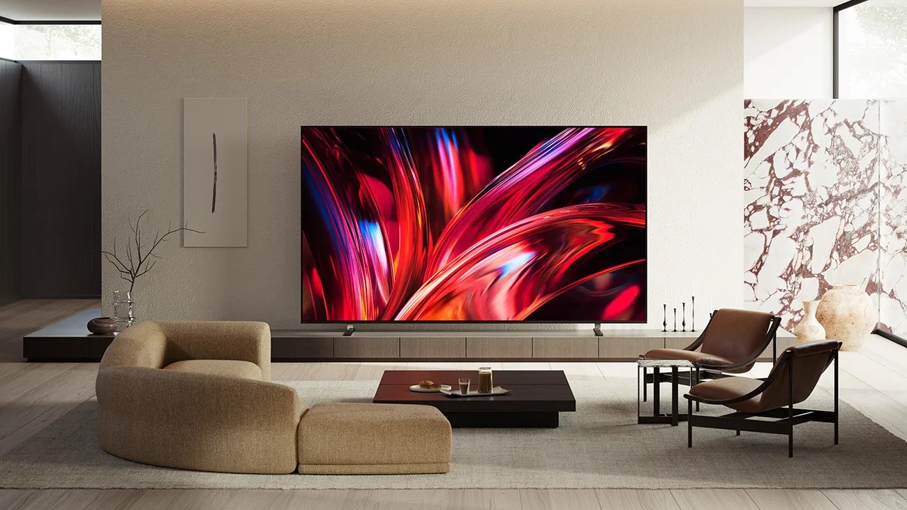 Modern living room with a large flat-screen TV displaying colorful abstract art, beige sectional sofa, and two brown leather chairs.