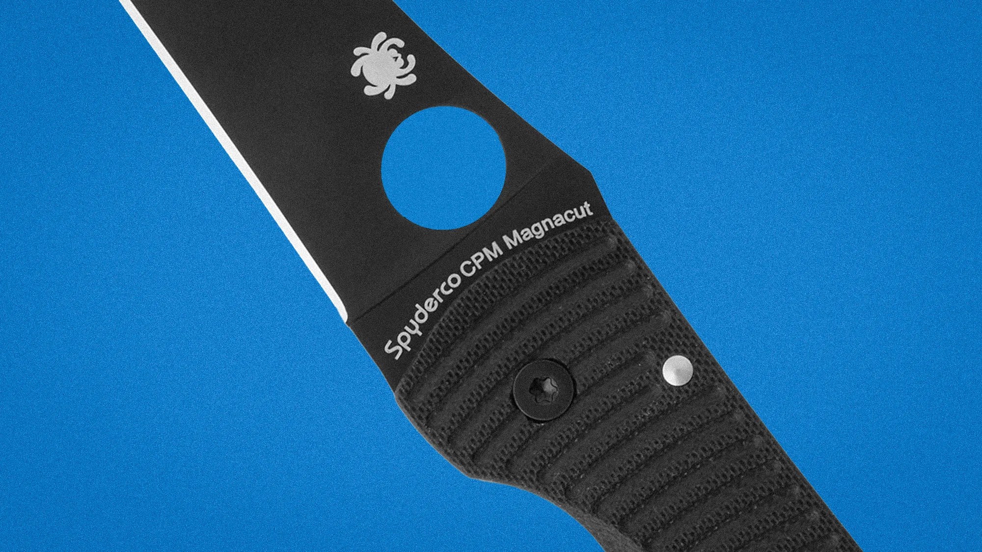 Close-up of a black Spyderco CPM Magnacut knife blade and textured handle against a blue background.