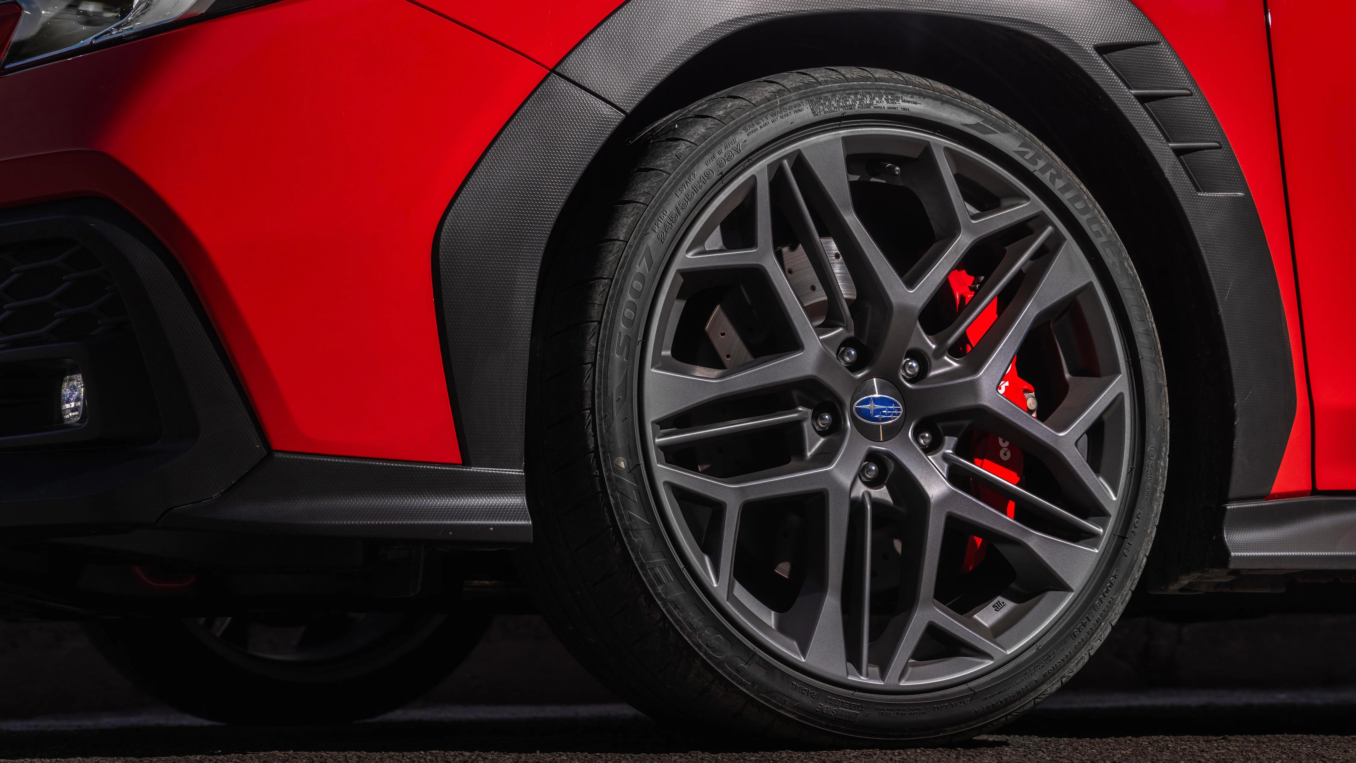 Close-up of a dark gray Subaru alloy wheel with a red brake caliper on a red car.