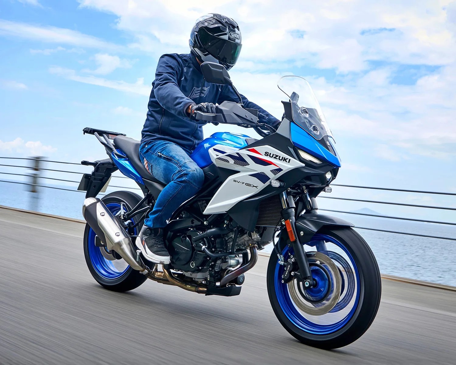 Blue and white Suzuki SV7 GX motorcycle with blue wheels ridden on a coastal road.