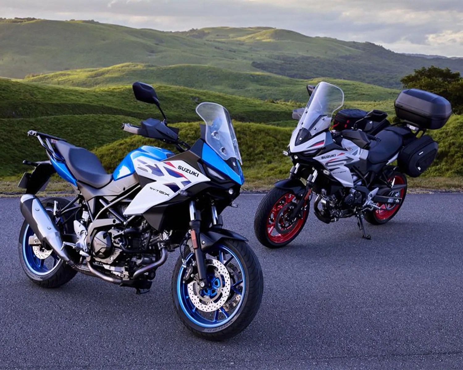 Two Suzuki sport touring motorcycles, one blue and white with blue wheels and one white and black with red wheels and luggage boxes, parked on a road with green hills in the background.