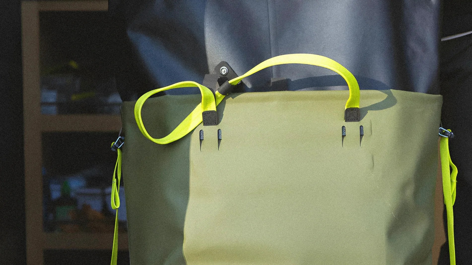 Olive green tote bag with bright yellow handles and shoulder strap worn by a person in a dark outfit.
