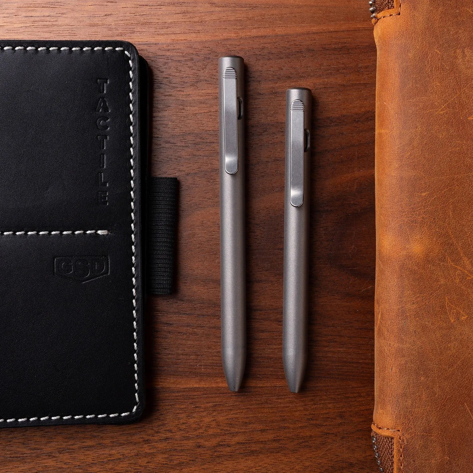 Two matte silver metal pens with clips placed vertically on a wooden surface between a black stitched leather notebook and a brown leather case.