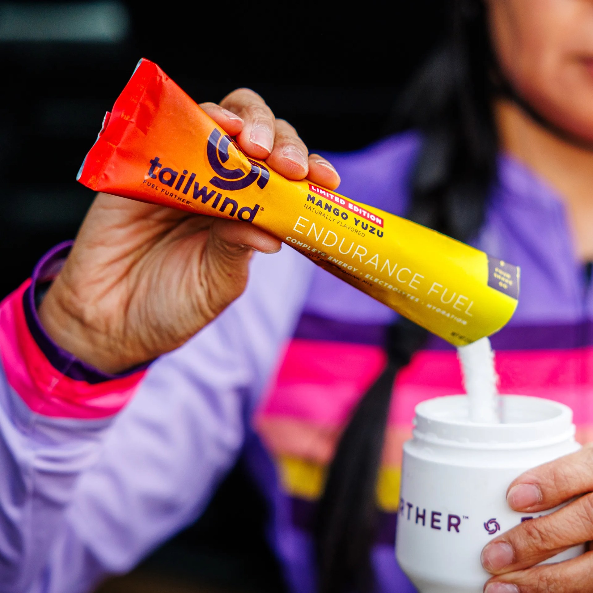 Hand holding a Tailwind Endurance Fuel Mango Yuzu packet pouring powder into a white bottle.
