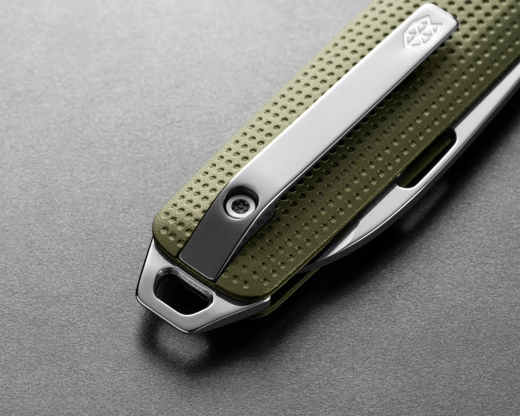 Close-up of a green textured folding knife handle with a silver metal clip and screws on a gray surface.