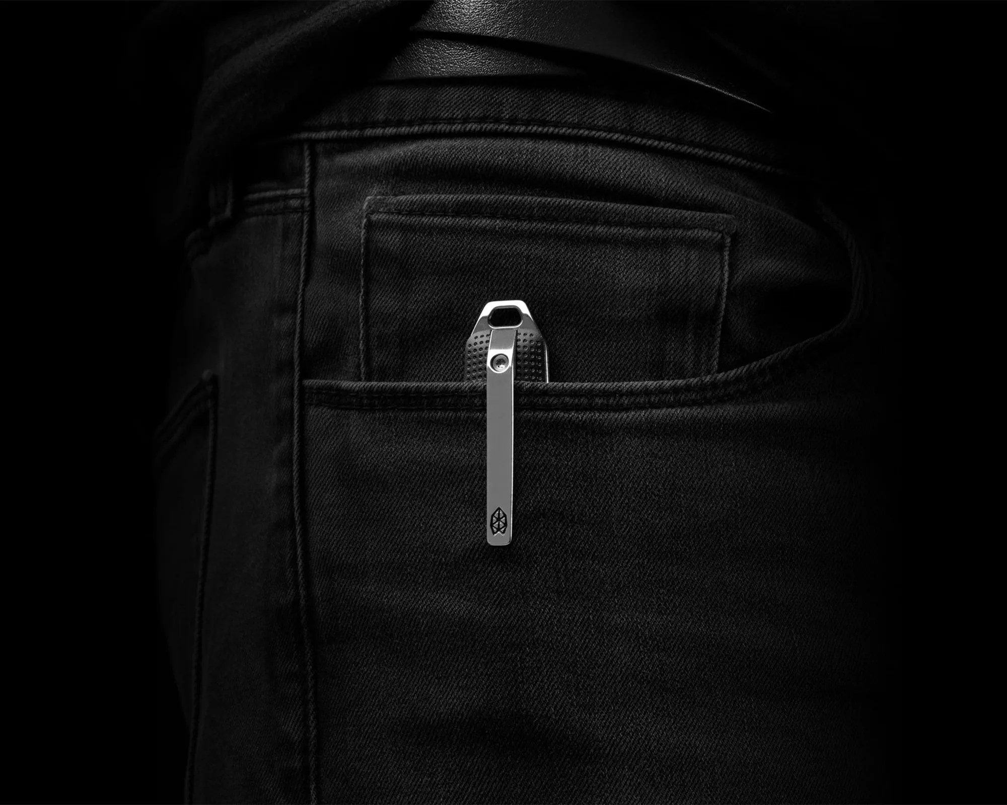 Silver metal clip with a textured grip attached to the back pocket of black denim jeans.