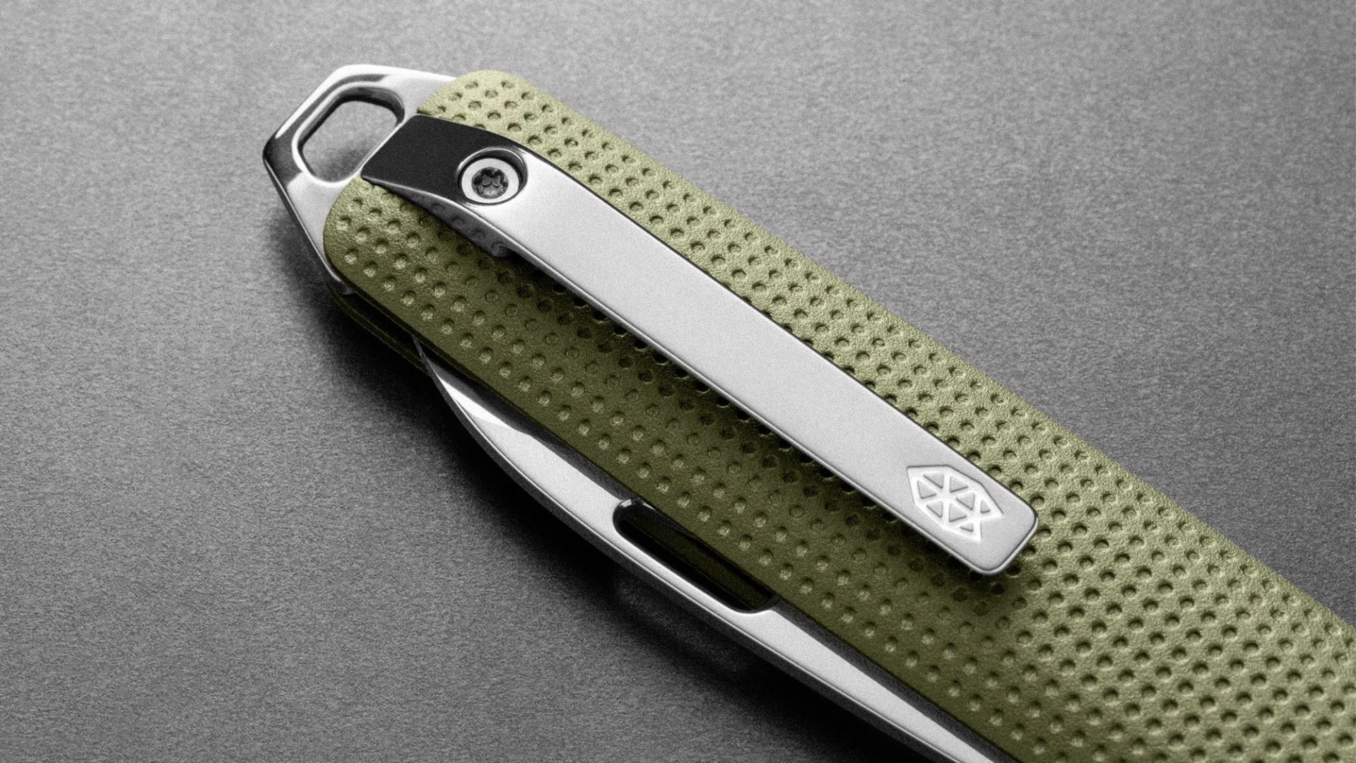 Close-up of a green textured folding knife handle with a silver metal clip and a small emblem on a gray surface.