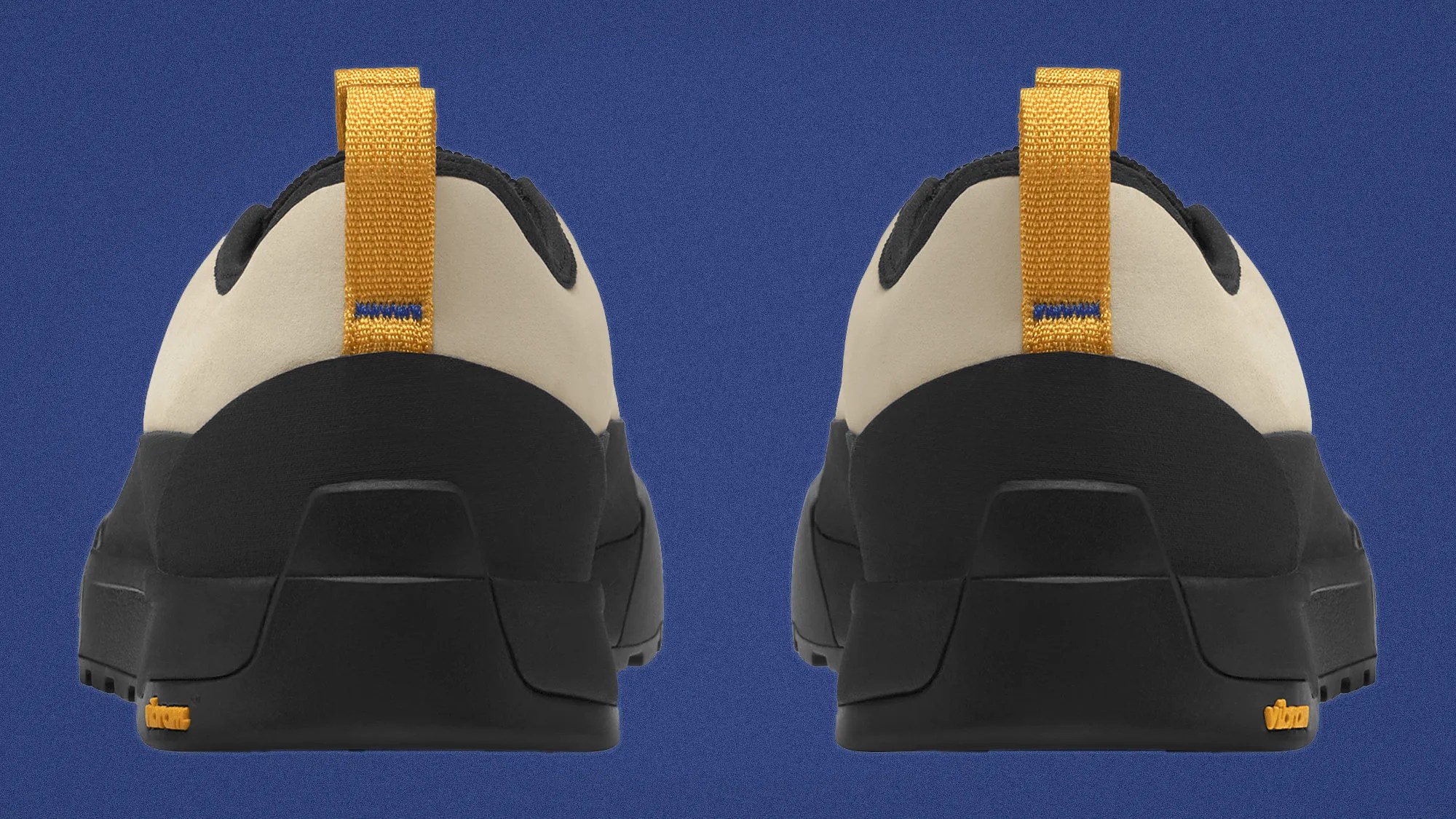 Rear view of beige and black shoes with yellow pull tabs and Vibram logo on the sole against a blue background.