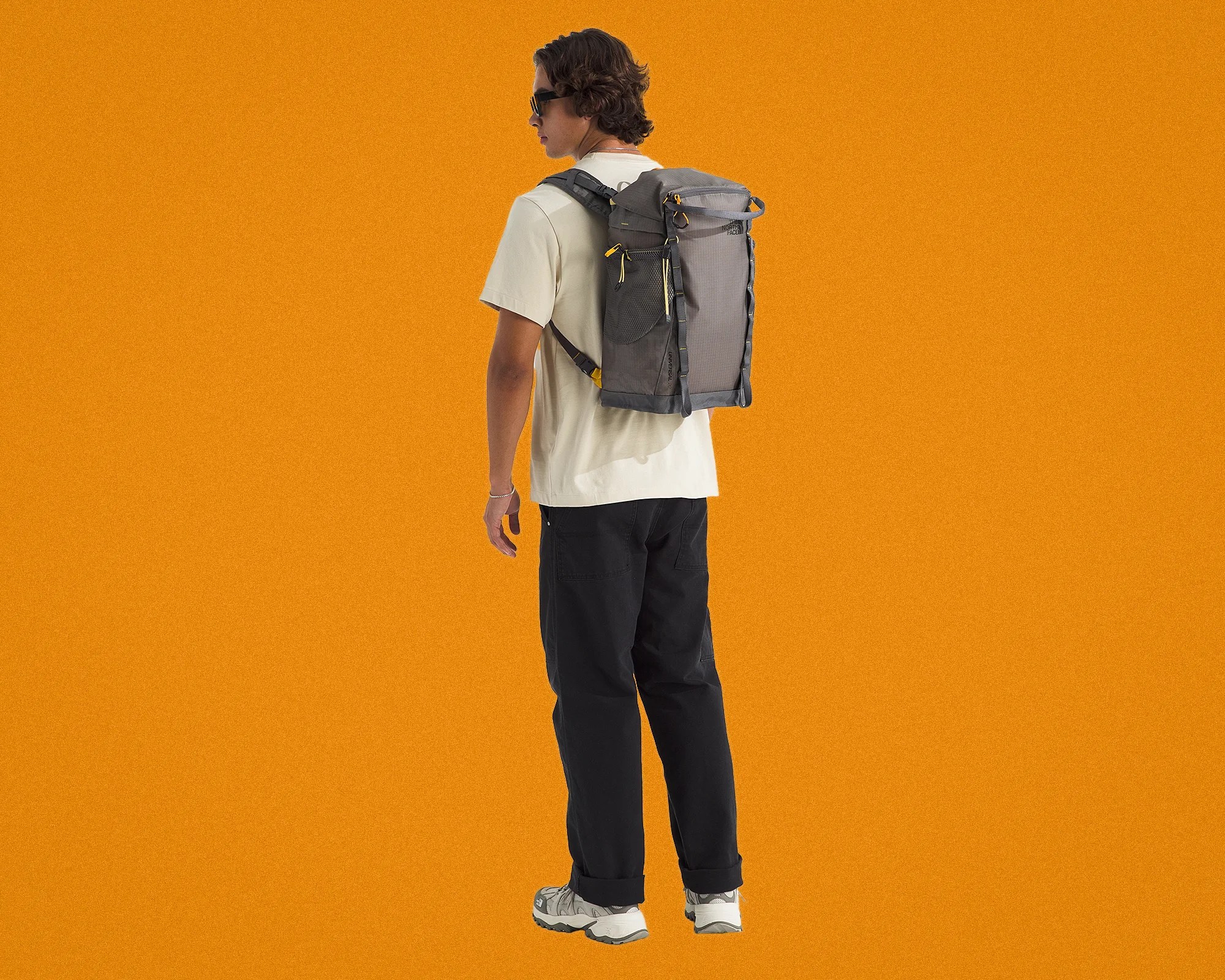 Person wearing a gray North Face backpack with yellow zipper pulls, black pants, and white sneakers against an orange background.