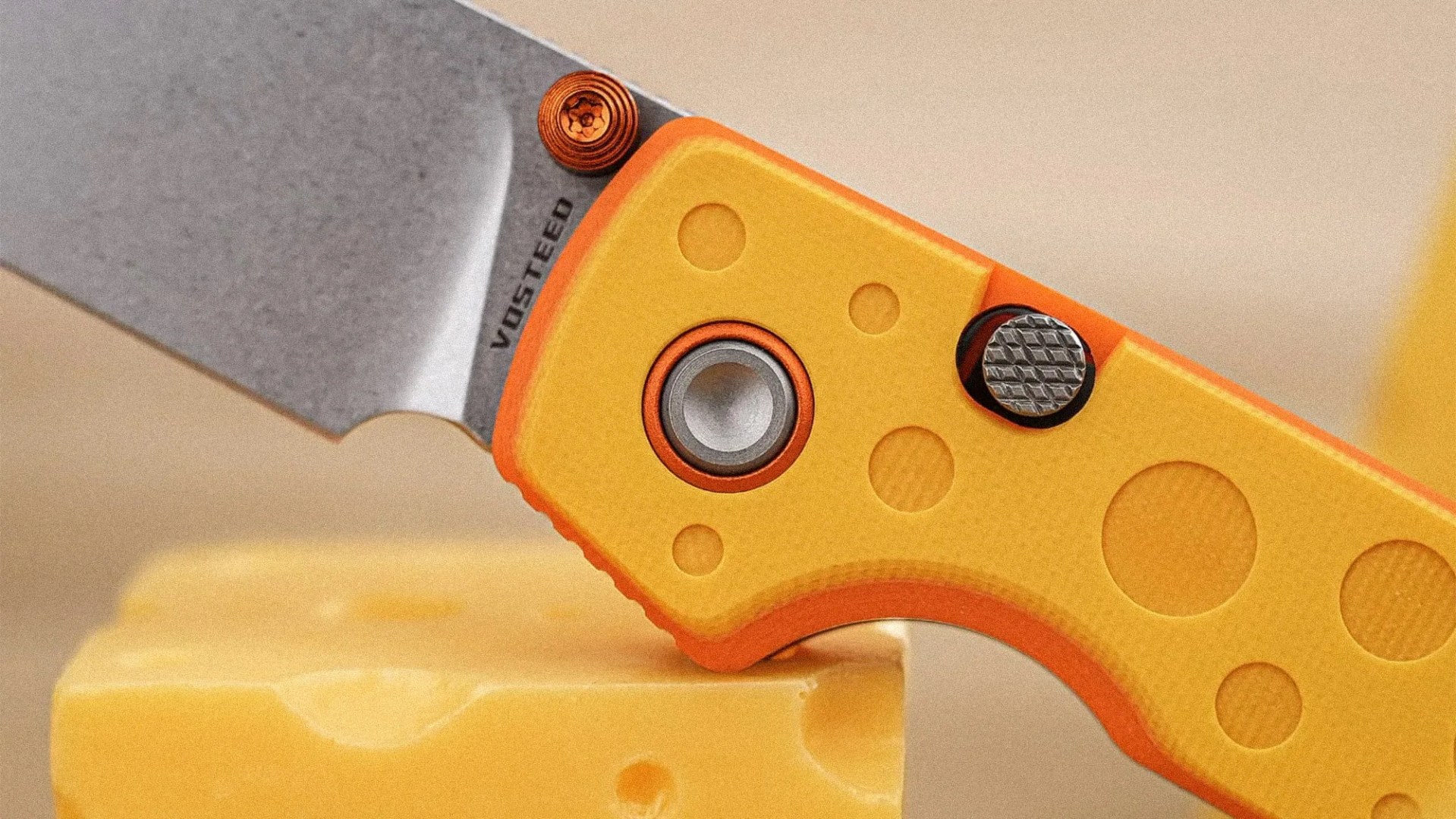 Close-up of a yellow and orange Vosteed folding knife blade cutting a block of Swiss cheese.